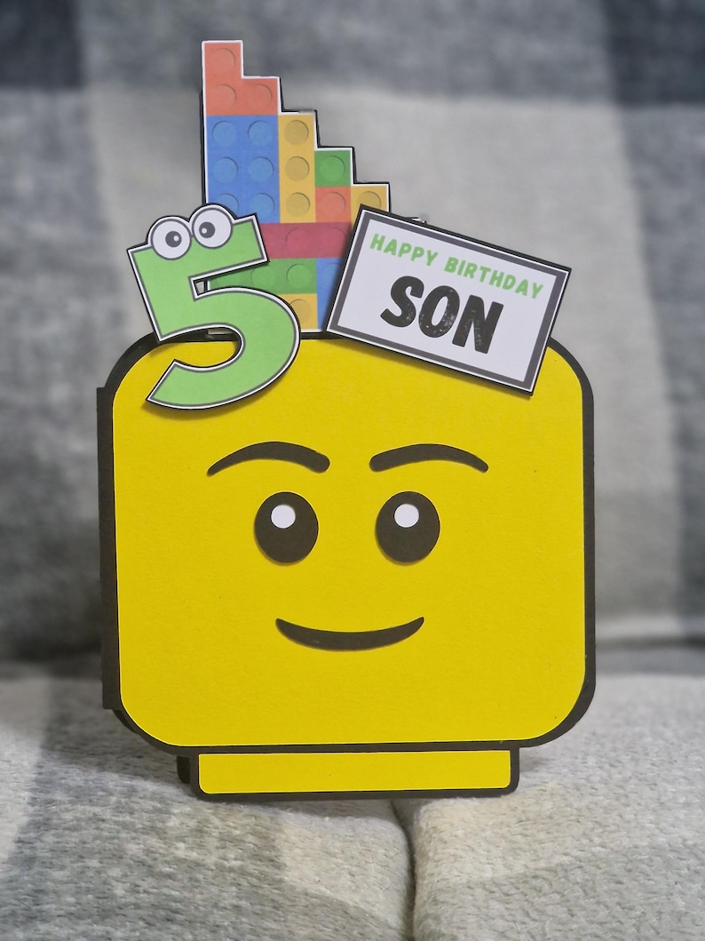 May include: A yellow birthday card in the shape of a Lego head, featuring a green number 5 and a sign reading "Happy Birthday Son." The card is adorned with colourful Lego bricks.