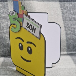 May include: A yellow birthday card in the shape of a Lego figure's head, featuring a green number 5 and a sign saying "HAPPY BIRTHDAY SON." The card is decorated with a stack of colourful Lego bricks.