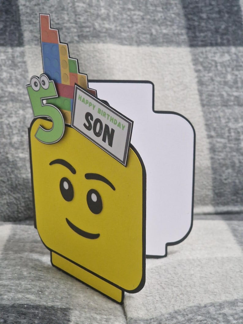 May include: A yellow birthday card in the shape of a Lego figure's head, featuring a green number 5 and a sign saying "HAPPY BIRTHDAY SON." The card is decorated with a stack of colourful Lego bricks.