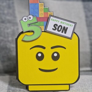 May include: A yellow Lego-style birthday card featuring a smiling face. A green number 5 with googly eyes is on the left, and a stack of colourful Lego bricks is on top. The card reads "HAPPY BIRTHDAY SON" on a white rectangle.