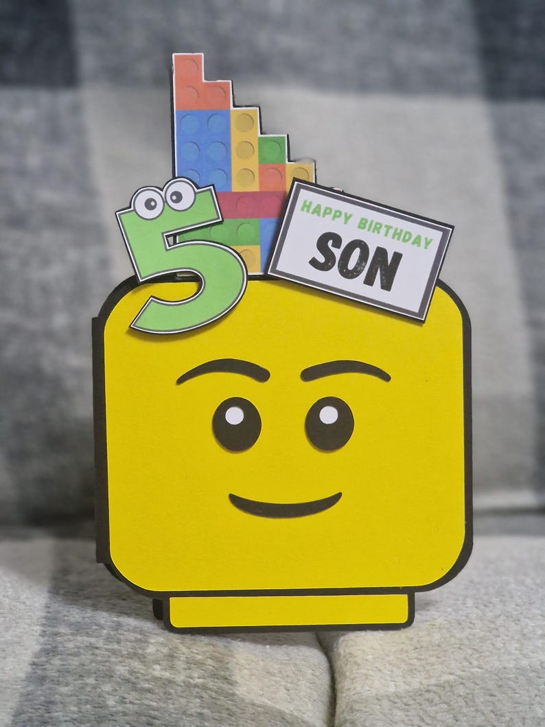 May include: A yellow Lego-style birthday card featuring a smiling face. A green number 5 with googly eyes is on the left, and a stack of colourful Lego bricks is on top. The card reads "HAPPY BIRTHDAY SON" on a white rectangle.
