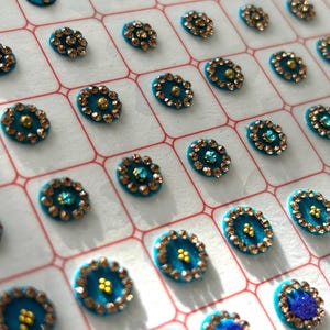 May include: A sheet of turquoise and gold decorative stickers. The stickers are arranged in rows and columns on a white background. Each sticker is a small, round, jeweled design.