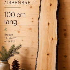 Può includere: Una tavola di legno chiaro con bordi naturali e nodi visibili. Il testo sull'immagine recita ZIRBENBRETT, 100 cm lang, e Breiten 14-26 cm erhältlich. La tavola presenta una superficie liscia e chiara con bordi in corteccia scura e ruvida.