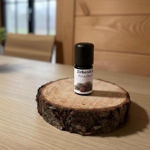 Könnte beinhalten: Eine kleine Glasflasche mit schwarzem Deckel, beschriftet mit "Zirbenöl Pinus Cembra", steht auf einem rustikalen, runden Holzuntersetzer. Das Etikett zeigt eine Illustration von Kiefernzweig und Zapfen. Der Untersetzer hat einen rauen Rand und eine hellbraune Oberfläche.