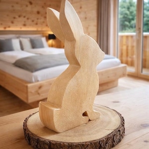 Pode incluir: Uma figura de coelho de madeira castanha clara, esculpida com linhas simples, assenta numa base redonda de madeira com uma borda de casca. O coelho tem orelhas compridas. A base tem cerca de 15 cm de diâmetro.
