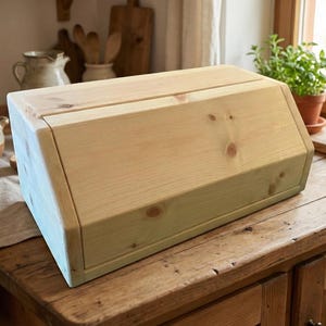 Könnte beinhalten: Eine hellfarbene Brotdose aus Holz mit einem Klappdeckel. Die Box ist rechteckig mit einer schrägen Vorderseite und steht auf einer Holz-Arbeitsplatte. Die Küchenszene umfasst Töpferwaren und Pflanzen.