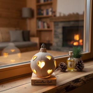 Op de afbeelding: Een houten, bolvormige lamp met hartvormige uitsparingen, van binnenuit verlicht. De lamp staat op een houten voet met de tekst "Gesund mit Zirbe". Daarnaast dennenappels, een kleine fles en een plant, alles op een houten oppervlak.
