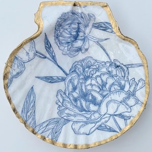 May include: A decorative scallop shell dish featuring a white surface with a blue ink-style floral pattern of peonies and leaves. The scalloped edges of the shell are finished with a metallic gold trim that follows the natural contour of the shell.