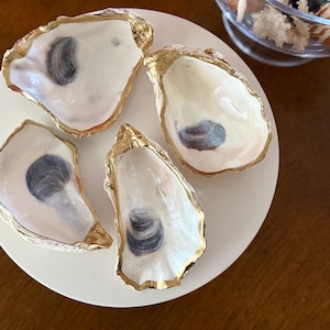 May include: Four oyster shells with white interiors and dark gray muscle scars, each featuring a metallic gold painted rim. The shells are arranged on a circular white surface, showcasing their natural concave shapes and smooth, glossy finishes.