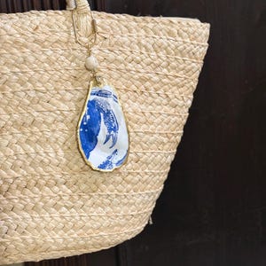 May include: A woven straw tote bag featuring a hanging decorative ornament made from a single oyster shell. The shell is painted with a blue and white crab design, finished with a gold-toned metallic edge, a small white bead, and a gold clasp.