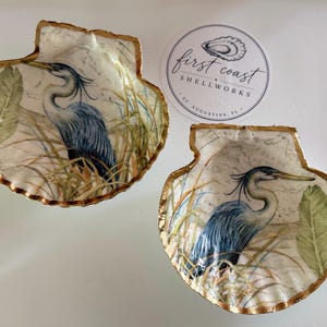 May include: Two decorative scallop shells featuring a blue heron illustration amidst tall grasses and green leaves. The edges of the shells are painted with a metallic gold finish. A circular white sticker with the text First Coast Shellworks is included.