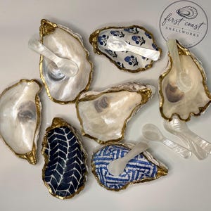 May include: A collection of seven oyster shell dishes with gold-painted edges, some featuring blue and white patterns, accompanied by small white mother-of-pearl spoons. A circular sticker with the text First Coast Shellworks is visible.