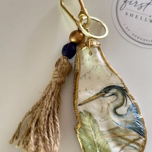 May include: A decorative keychain featuring a decoupage oyster shell with a blue heron and green leaf illustration, finished with a gold-painted edge. The shell hangs from a gold-tone metal clasp with a jute tassel, a dark blue bead, and a gold bead.