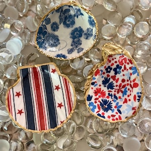 May include: Three decorative seashells with gold-painted edges. One shell features a blue and white floral pattern, another has red and blue vertical stripes with red stars, and the third displays a red and blue floral print on a white background.