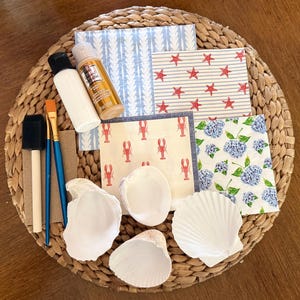 May include: A craft kit containing four white seashells, two paintbrushes, a black foam brush, a bottle of metallic gold acrylic paint, a bottle of white paint, and four decorative paper napkins featuring starfish, lobster, and hydrangea patterns.