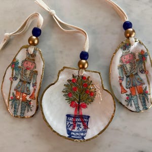 May include: Three decorative ornaments crafted from natural seashells with gold-painted edges. Two shells feature hand-painted nutcracker figures in blue and orange uniforms, while the center shell displays a potted topiary with a red bow.