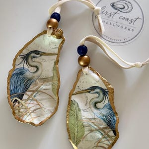 May include: Two decorative oyster shell ornaments featuring a blue heron illustration on a map background. Each shell has gold-painted edges, a cream ribbon, and a blue and gold bead. A circular sticker reads First Coast Shellworks Augustine, FL.