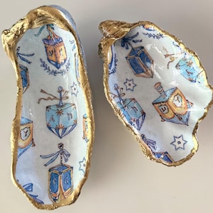 May include: Two decorative oyster shells with gold-painted edges. The interior of each shell features a white background with a repeating pattern of blue and orange dreidels, small blue stars, and delicate blue ribbon accents throughout.