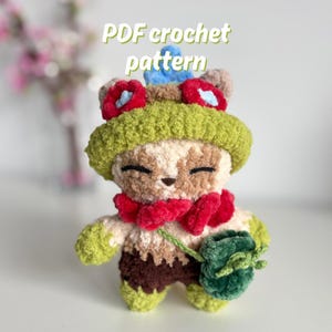 Könnte beinhalten: Handgefertigte Häkelpuppe mit einem skurrilen Design. Die Puppe hat einen grünen Hut, rote Akzente und einen kleinen grünen Beutel. Der Text "PDF crochet pattern" ist oben zu sehen. Die Puppe ist aus weichem, strukturiertem Garn gefertigt.