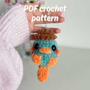 May include: A teal and orange crochet platypus keychain with a brown hat. The keychain has large black eyes and a small orange beak. The text "PDF crochet pattern" is in white. A handmade item.