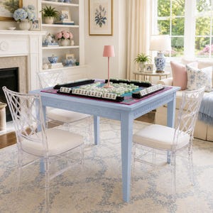 May include: A light blue square game table with a Greek key pattern on the apron, surrounded by four clear acrylic chairs with white seat cushions. A mahjong set is arranged on a colorful mat on the table, topped with a small pink lamp.