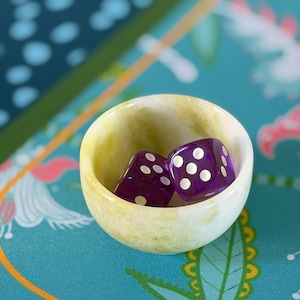 May include: A small, round, pale yellow and white stone bowl containing two translucent purple six-sided dice. The dice feature white circular pips on each face, with one die showing five pips and the other showing one pip clearly visible.