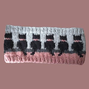 May include: A knitted headband featuring a repeating pattern of black cat silhouettes against a light gray background. The top and bottom edges are finished with a ribbed texture, and the lower section includes a solid dusty pink horizontal band.