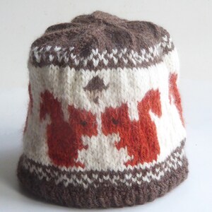 Può includere: Un cappello di lana marrone e bianco con un motivo di scoiattoli e una piccola ghianda sulla cima. Gli scoiattoli sono arancioni e marroni.