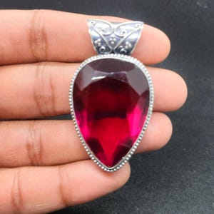 Könnte beinhalten: Ein tropfenförmiger Anhänger mit einem großen, facettierten, tiefroten Edelstein. Der Edelstein ist in einem silberfarbenen Metall mit einem dekorativen Bügel gefasst. Der Anhänger wird in einer Hand vor dunklem Hintergrund gehalten.