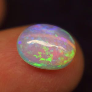 Può includere: Primo piano di una gemma di opale di forma ovale. L'opale mostra un vibrante gioco di colori, con lampi di tonalità verdi, rosa e blu. La gemma è tenuta tra due dita, su uno sfondo scuro.