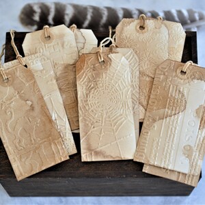 May include: A collection of vintage-style paper tags with embossed designs, including a spiderweb, floral patterns, and text. Each tag is a light brown color, strung with twine, and rests on a dark wooden box. The tags have a distressed, aged appearance.