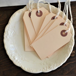 May include: A collection of blank, peach-colored price tags with brown eyelets and white string, arranged on a white, scalloped-edge plate. The plate sits on a dark wooden surface, creating a neutral backdrop for the tags.