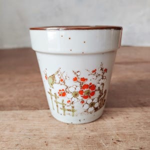 May include: A white ceramic cup with a brown rim featuring a hand-painted design of a small bird perched on a branch with red and white blossoms and a green fence. The exterior surface has subtle brown speckles scattered across the upper portion.