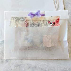 May include: A white paper package sealed with a decorative strip and a purple bow. The decorative strip features vintage-style floral and text designs. The package is likely for a gift or craft item.