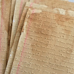 May include: A stack of aged paper sheets with embossed script and red vertical lines. The paper has a warm, brown tone with darker edges, suggesting an antique or vintage aesthetic. The embossed text adds a tactile element.