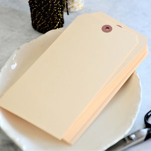 May include: A stack of blank, cream-colored gift tags with a brown eyelet hole. The tags are on a white plate with a pair of black scissors.