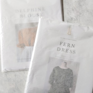 May include: Two white paper packages, secured with small gold clips, are displayed. The package on the left reads "DELPHINE BLOUSE" in a soft font. The package on the right reads "FERN DRESS" with "BY GALIA PATTERNS" above it. The packages contain sewing patterns.