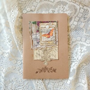 May include: A brown notebook with a collage of vintage ephemera. The collage includes a butterfly image, floral accents, and the text "Paris - 2 ar". A fairy illustration and floral stamp are also visible.