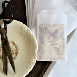 May include: A still life composition featuring a vintage aesthetic. A pair of antique scissors rests on a floral-decorated plate. A small paper bag with a butterfly stamp is visible, suggesting a craft or stationery theme.