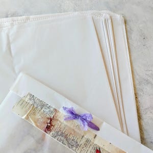 May include: A stack of white paper bags, with a decorative strip and a purple ribbon. The bags are rectangular and appear to be made of a thin, translucent material. The decorative strip has a vintage design.