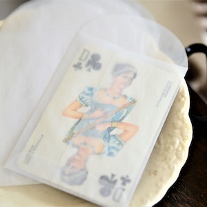 May include: A vintage playing card featuring a woman in a blue and white dress holding a staff. The card is a Queen of Clubs and has the text "La Belle Française" on the back.