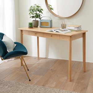 May include: A light oak desk with two drawers and brass handles. A blue radio, potted plant, and open book are on the desk. A teal chair with gold legs is in the foreground, and a round mirror hangs on the wall.