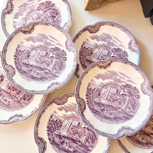 May include: A set of seven antique white and purple decorative plates. Each plate features a detailed landscape scene in the center, surrounded by a scalloped edge with a decorative border. The plates are arranged on a light surface.