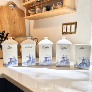 Op de afbeelding: Vijf witte keramische potten met blauwe en witte windmolenontwerpen. De potten zijn gelabeld met "Thee", "Ontbijtgranen", "Koffie", "Suiker" en "Rijst".