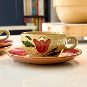 May include: A set of ceramic teacups and saucers. The cups and saucers are painted with a floral design featuring a red tulip, green leaves, and small white flowers. The cups have a light yellow background and a rounded handle. The saucers are a reddish-pink color.