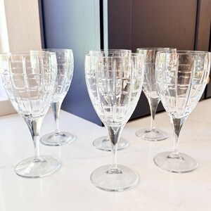 May include: Six clear crystal wine glasses with a grid pattern on the bowl. The glasses have long stems and round bases. They are arranged on a white countertop near a window and dark cabinets.