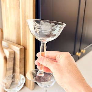 Könnte beinhalten: Ein klares Coupé-Cocktailglas mit einem zarten, geätzten Blumenmuster am Rand. Der Stiel ist glatt und führt zu einem runden Fuß, wobei das Glas eine breite, flache Schalenform aufweist, die sich zum Servieren verschiedener Getränke eignet.