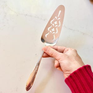 May include: A silver pie server with a decorative openwork design on the blade. The handle has an ornate floral pattern. The server is held against a white marble surface.