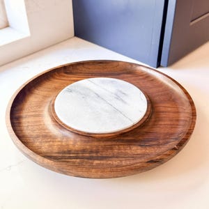 May include: A circular serving tray crafted from dark brown wood with a prominent, smooth, white marble inlay centered in the middle. The wooden base features a visible grain pattern and a raised outer rim, creating a contrast with the stone.