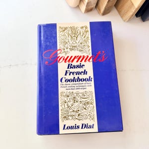 May include: A hardcover cookbook titled Gourmet's Basic French Cookbook by Louis Diat. The cover is deep blue with a central cream-colored vertical band featuring gold line illustrations of food and wine, and red and black text.